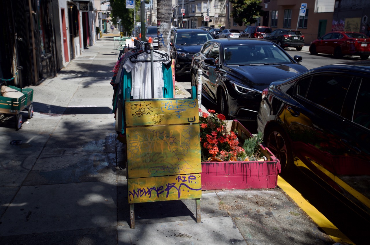 A city sidewalk scene showing a graffiti-covered yellow sidewalk sign, a row of parked cars, potted red flowers, and clothes hanging on racks under partial sunlight and shade.