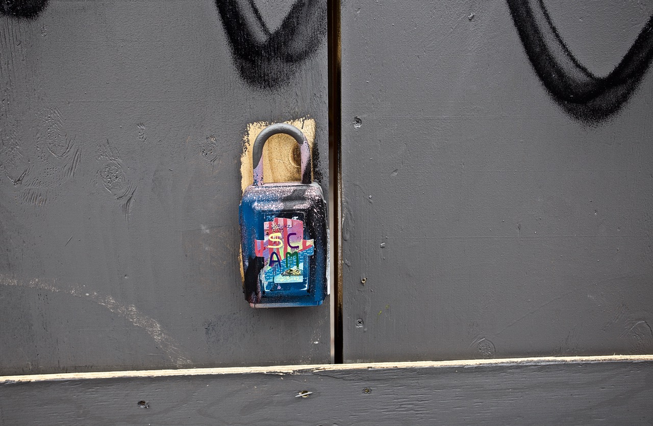 A padlock attached to a grey wooden door is painted with colorful graffiti and the word SCAM.
