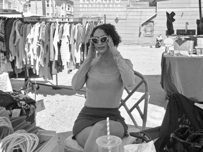 A woman sits at an outdoor market trying on sunglasses. Clothing racks and a "Mexican Cooking" sign are visible in the background. A drink with a straw is placed in the foreground.