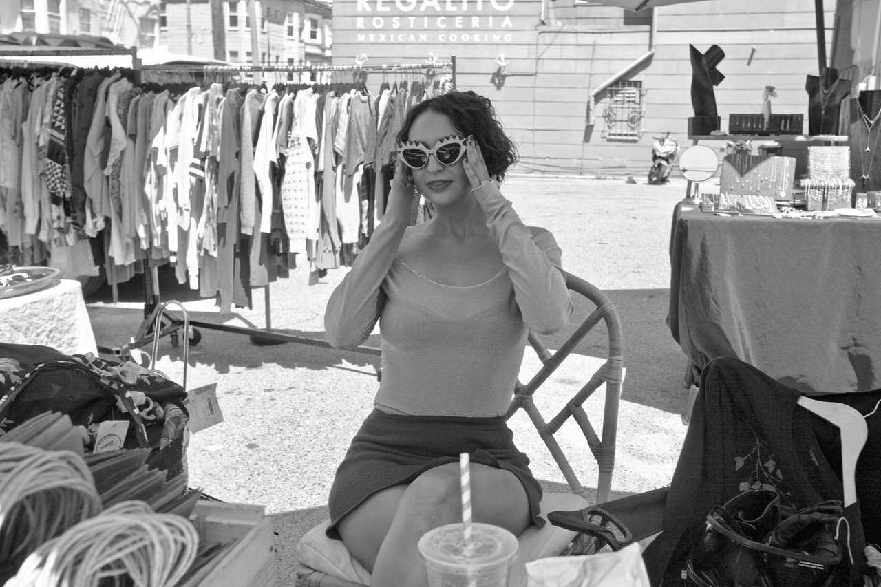 A woman sits at an outdoor market trying on sunglasses. Clothing racks and a "Mexican Cooking" sign are visible in the background. A drink with a straw is placed in the foreground.