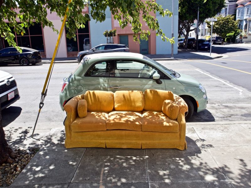 A yellow couch is placed on a sidewalk next to a parked car on an urban street with buildings and trees in the background.