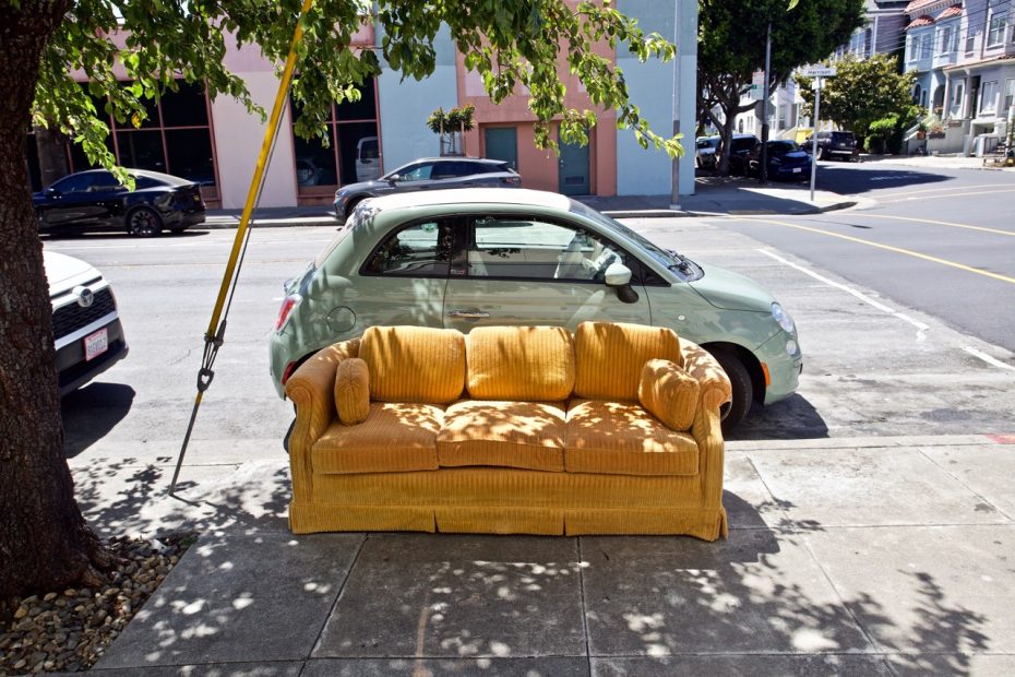 A yellow couch is placed on a sidewalk next to a parked car on an urban street with buildings and trees in the background.