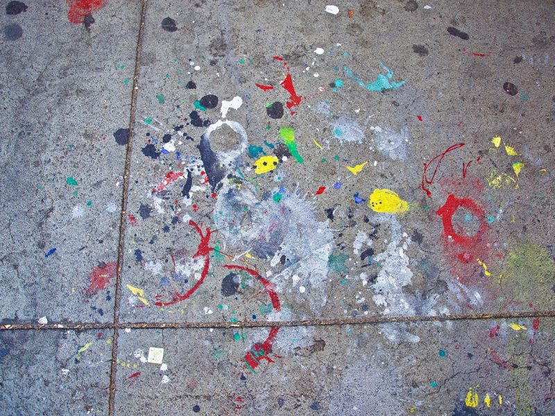 A concrete sidewalk stained with various colors of paint splatters and splotches, displaying a chaotic mix of red, yellow, green, blue, and black marks.