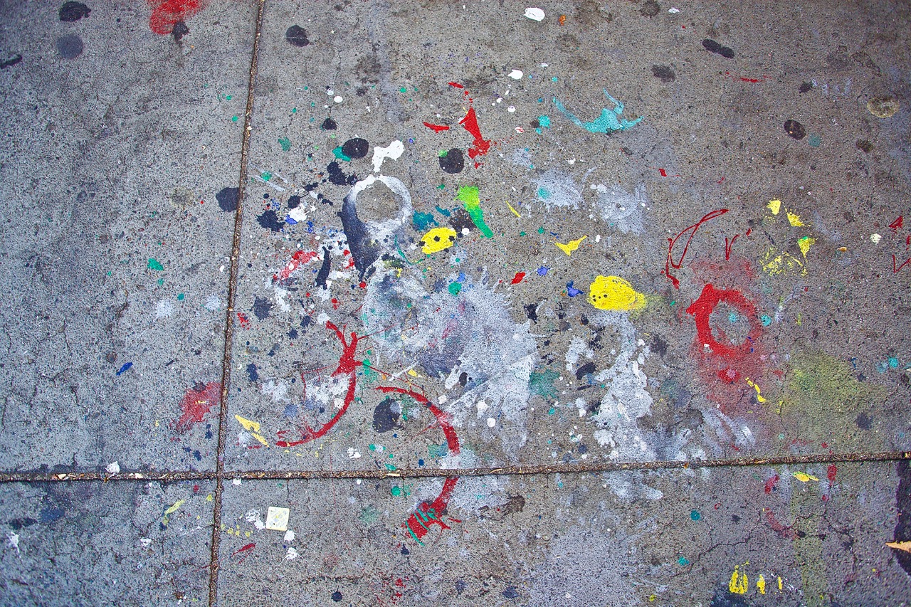 A concrete sidewalk stained with various colors of paint splatters and splotches, displaying a chaotic mix of red, yellow, green, blue, and black marks.