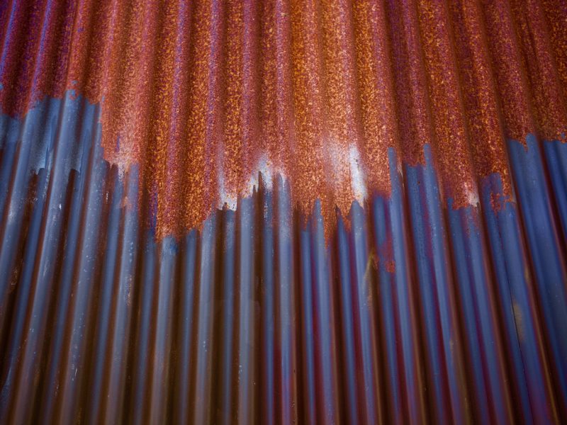 Close-up of a corrugated metal sheet showing rust formation at the top and blue paint or primer peeling off towards the bottom.