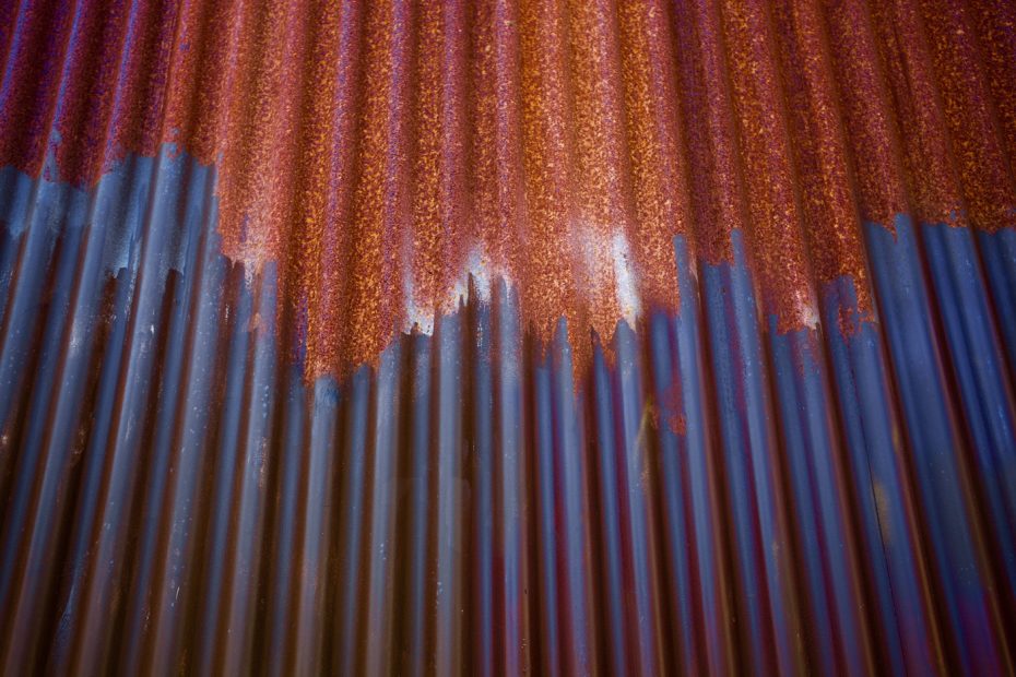 Close-up of a corrugated metal sheet showing rust formation at the top and blue paint or primer peeling off towards the bottom.