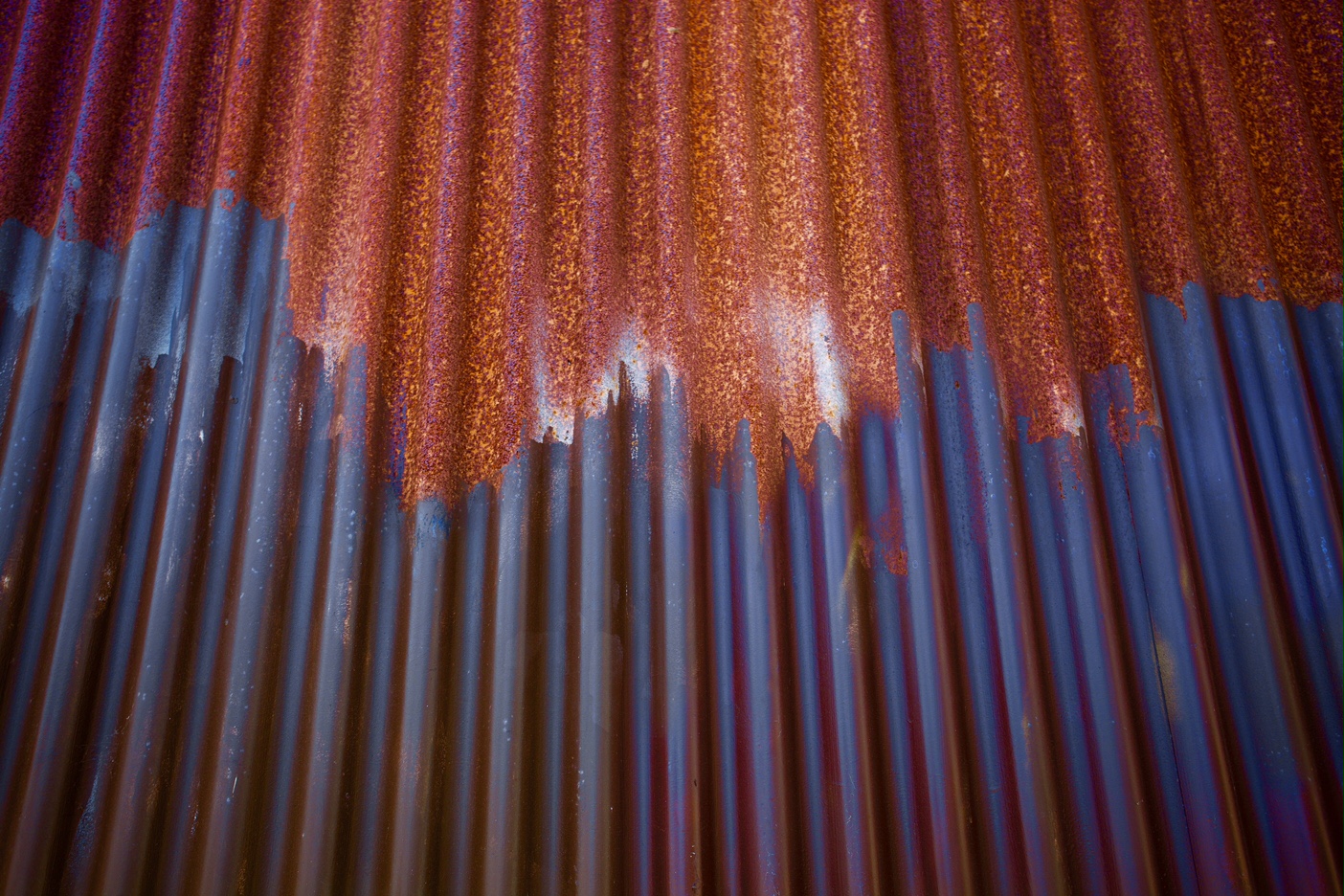 Close-up of a corrugated metal sheet showing rust formation at the top and blue paint or primer peeling off towards the bottom.