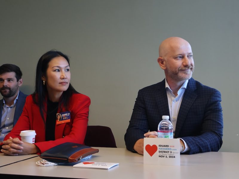 Three individuals sit at a table with papers, water bottles, and a small sign. The woman in the center wears a red blazer and holds a cup. The man on the right has a tablet displaying the "District 3" text.