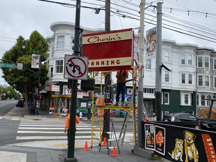 A person stands on scaffolding, working on a sign that reads "Cherin's PARKING" at a street corner with a crosswalk and traffic sign. Other signage and buildings are in the background.