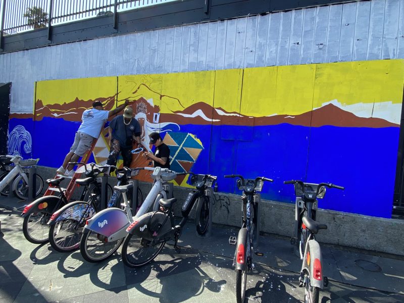 Three people are painting a mural on a wall behind a row of parked bikes. The mural features mountains, a sunset, and geometric shapes.