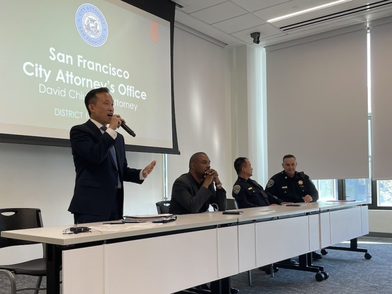 Four officials are seated at a table in a conference room while one of them speaks into a microphone, with a presentation slide behind them titled "San Francisco City Attorney's Office.