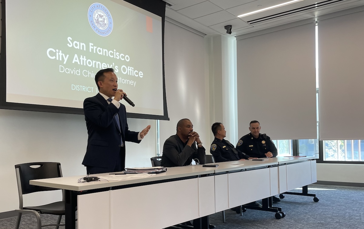 Four officials are seated at a table in a conference room while one of them speaks into a microphone, with a presentation slide behind them titled "San Francisco City Attorney's Office.