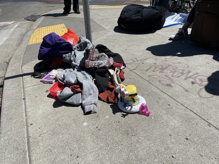 Assorted clothes, a stuffed toy, and personal items scattered on a sidewalk next to a pole, with people and luggage partially visible in the background. During a homeless sweep.