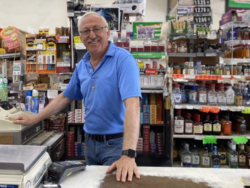 A man in a blue polo shirt stands behind the counter of a convenience store, smiling. Shelves stocked with various products, including liquor, cigarettes, and other items, are visible in the background.