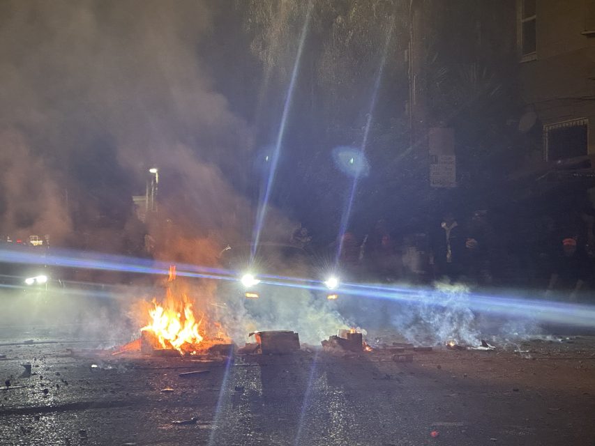 A street scene at night with a small fire burning amidst debris. Headlights from a vehicle shine through the smoke, illuminating the area. Several figures are faintly visible in the background, reminiscent of a chaotic July 4 celebration gone awry.