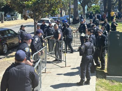A group of police officers in uniform set up metal barricades on a sidewalk in a park area, preparing for the eagerly anticipated hill bomb. Trees and cars are visible in the background, adding to the scenic setting.