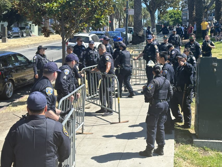 A group of police officers in uniform set up metal barricades on a sidewalk in a park area, preparing for the eagerly anticipated hill bomb. Trees and cars are visible in the background, adding to the scenic setting.
