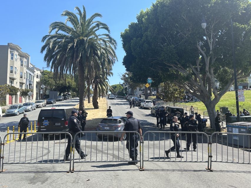 Police officers stand behind metal barricades on a street with a large palm tree and other trees around, with buildings on both sides of the street, prepared for any disruptions as skateboarders gather for an anticipated hill bomb.
