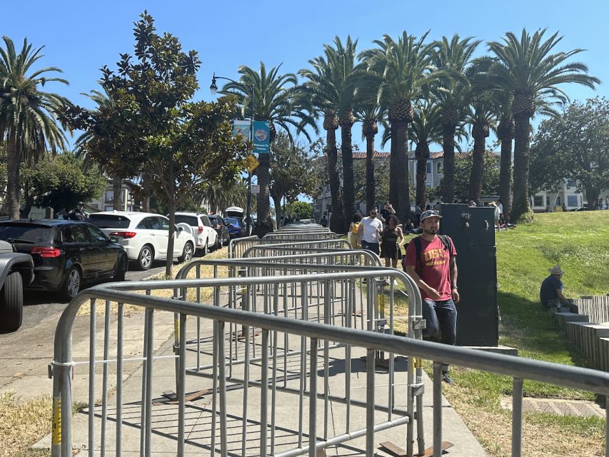 A series of metal barricades lines a path next to parked cars and a green space with palm trees. People are walking and standing on the grass and sidewalk, enjoying the day as skateboarders gather for a thrilling hill bomb nearby.