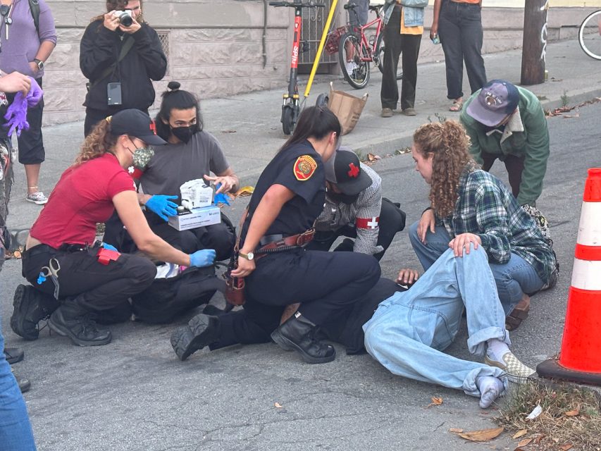 Two emergency responders assist an individual lying on the ground while another person helps. A bystander takes a photo, and a few others observe the scene nearby.