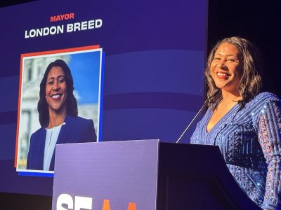 SF Mayor London Breed in a blue dress stands at a podium smiling, with an image and text on a screen behind her that reads "Mayor London Breed.