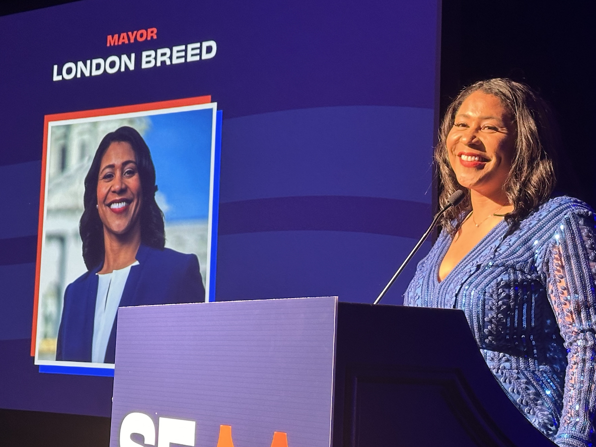 SF Mayor London Breed in a blue dress stands at a podium smiling, with an image and text on a screen behind her that reads "Mayor London Breed.
