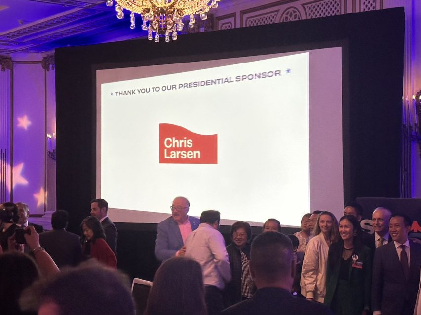 在一次活动中，一群人站在大屏幕前。屏幕上显示的是对赞助商Chris Larsen 的感谢致辞。会场是一个装饰一新的室内场所，吊灯熠熠生辉。布里德市长发表讲话。