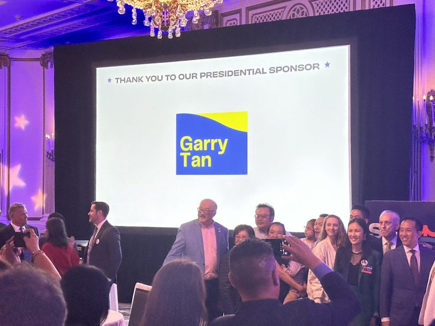 A group of people stands in front of a screen displaying a sponsorship message for Garry Tan at an event with chandeliers and ornate decor.