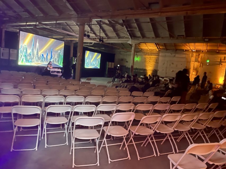 An indoor event space with rows of empty white chairs facing two large screens displaying a cityscape. A few people are scattered around the room.