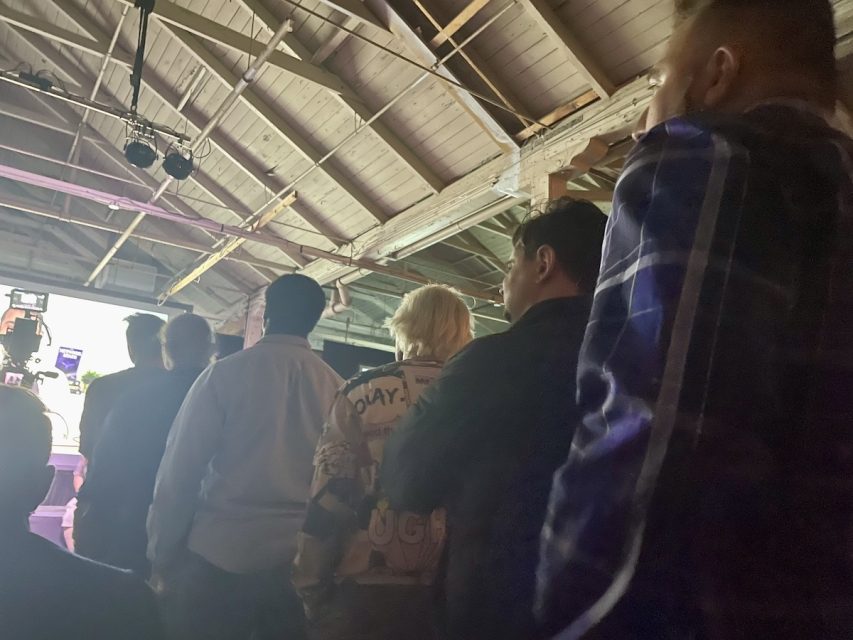A group of people stands under a wooden ceiling, facing away towards a bright screen or stage in a dimly lit room. Some of the individuals wear casual clothes while others are in suits.