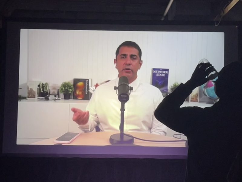 A person is speaking into a microphone in a studio-like setting, visible on a large screen. In the foreground, another person is drinking from a bottle.