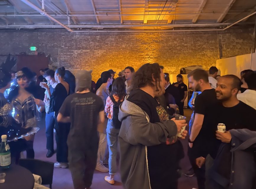 A group of people socializing in a dimly lit room at a party. Some are holding drinks and engaged in conversations. The background features exposed brick walls and industrial lighting.