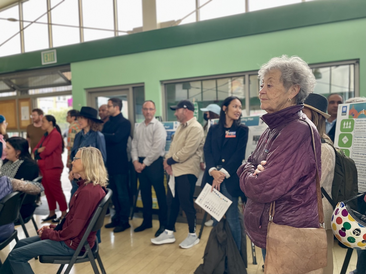 A group of people, including an elderly woman in a purple jacket, stand and sit indoors, attentively listening to something. Some are holding papers. A green wall and large windows are in the background.
