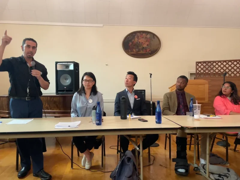 五人坐在小组讨论会上。左侧一人站着对着麦克风讲话，其他人在倾听。背景是一个扬声器和装饰墙。