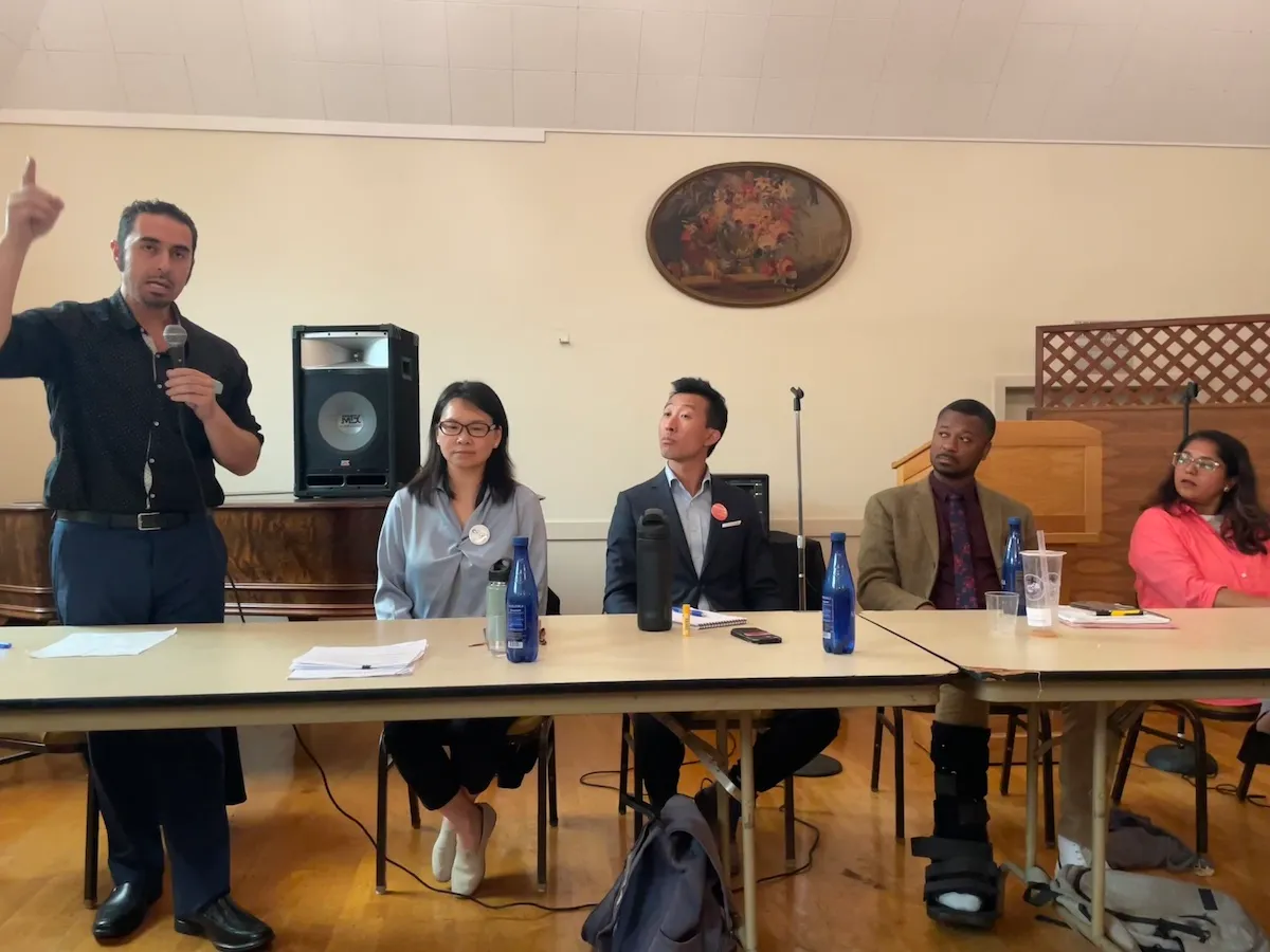 五人坐在小组讨论会上。左侧一人站着对着麦克风讲话，其他人在倾听。背景是一个扬声器和装饰墙。