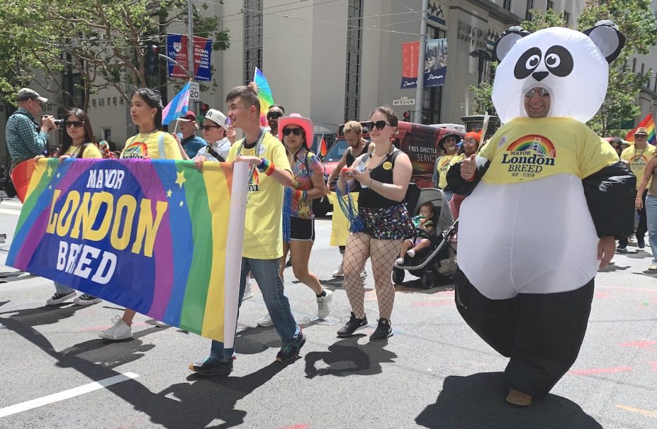 People march in a parade wearing yellow "Mayor London Breed" shirts, with one person in a panda costume and colorful flags. They carry a "Mayor London Breed" banner with a rainbow design.