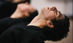 Close-up of a person lying on their back with their head tilted back and eyes closed, appearing relaxed. Another person is blurred in the background in a similar position.