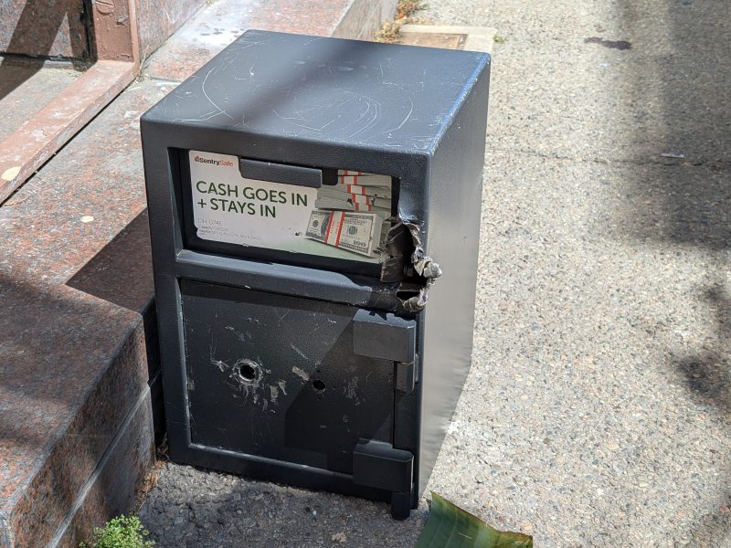 A black cash vault with visible damage, including holes and dents, stands on a concrete surface. A sign on it reads "CASH GOES IN + STAYS IN," but it appears to have been tampered with.