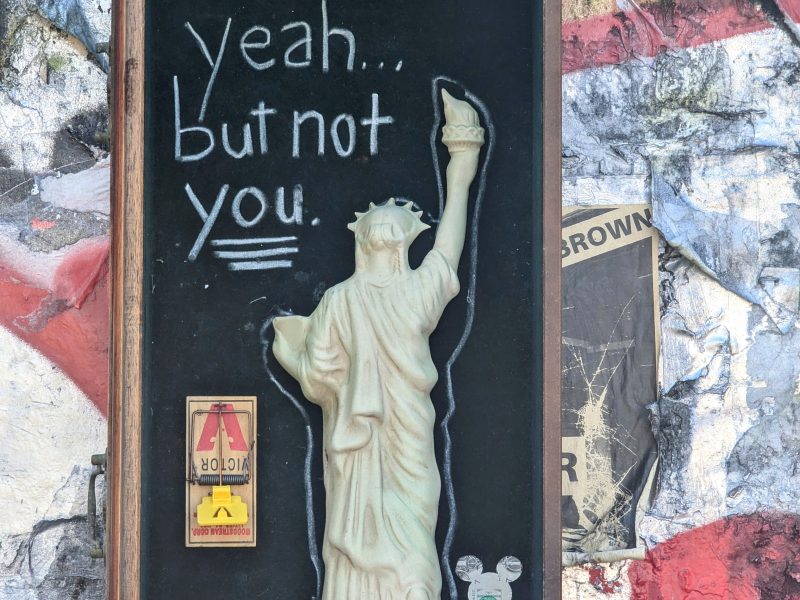 Street art featuring a framed image of the Statue of Liberty with a raised arm. Text above the statue reads "yeah... but not you." Collaged paper background with a mousetrap and stickers.