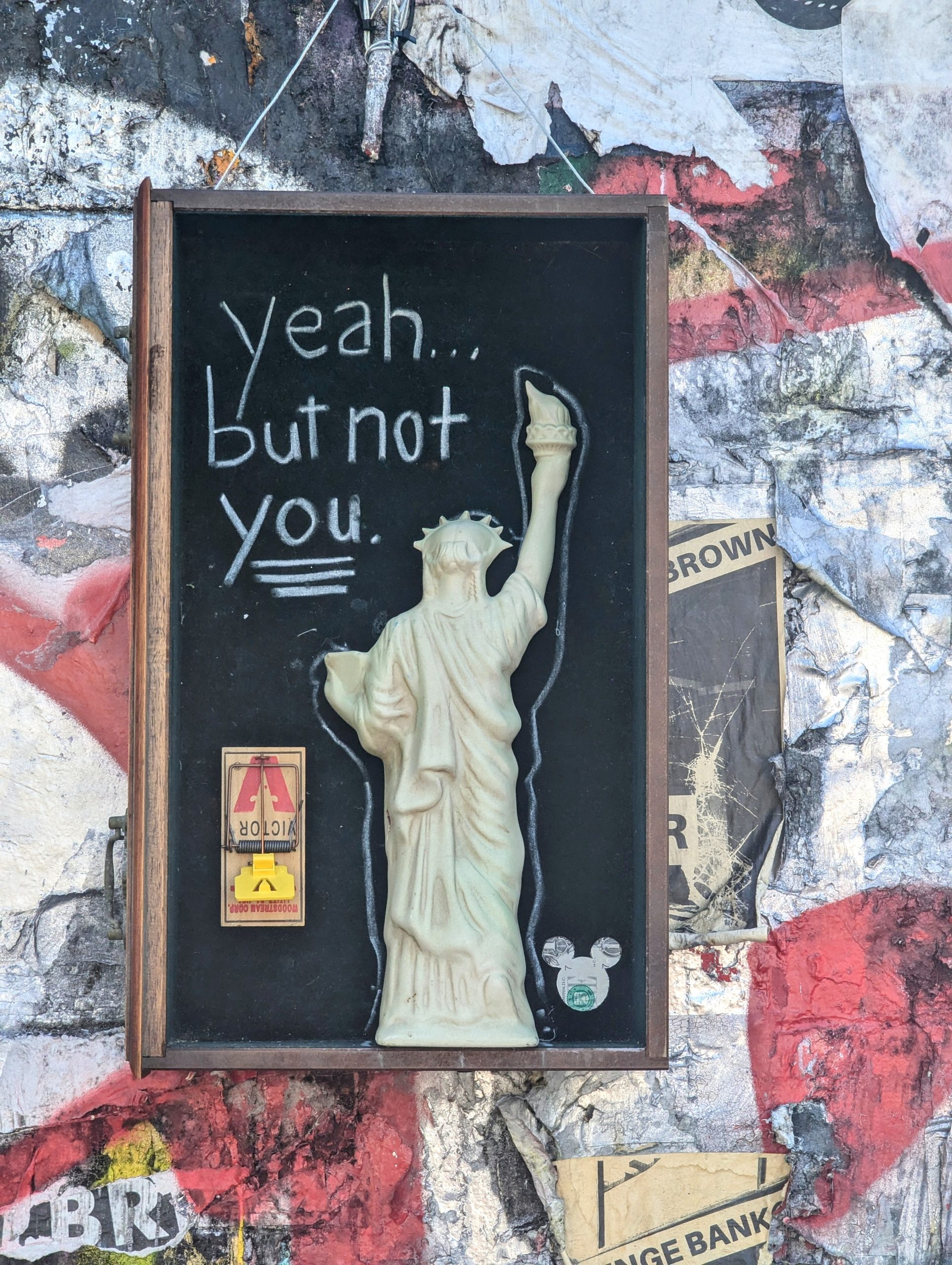 Street art featuring a framed image of the Statue of Liberty with a raised arm. Text above the statue reads "yeah... but not you." Collaged paper background with a mousetrap and stickers.