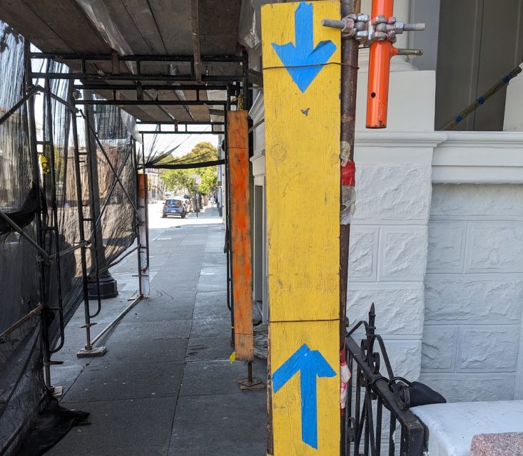 A yellow post with blue directional arrows stands on a sidewalk beside a building under construction. Scaffolding and protective netting line the rest of the sidewalk.