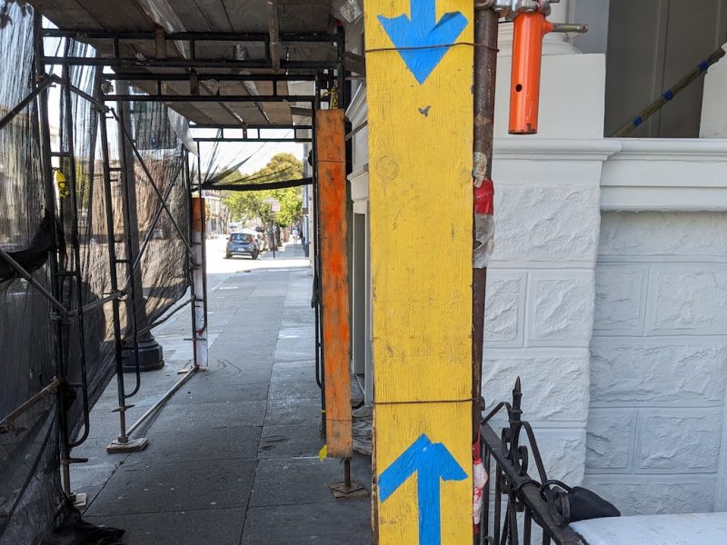 A yellow post with blue directional arrows stands on a sidewalk beside a building under construction. Scaffolding and protective netting line the rest of the sidewalk.