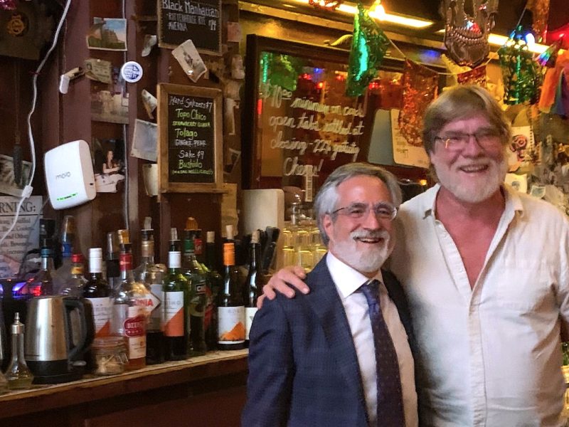 Two smiling men stand side by side at a busy bar, with bottles and decorations visible in the background.