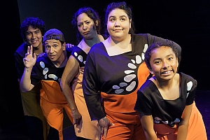 Five people in matching costumes pose together on stage, with four standing and one kneeling at the front. They are lit by stage lights against a black background.