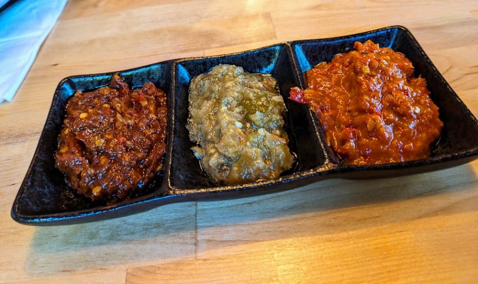 Three types of salsa in a black, divided serving dish on a wooden table: a red, chunky salsa on the left, a green, smooth salsa in the middle, and a red, thick salsa on the right.