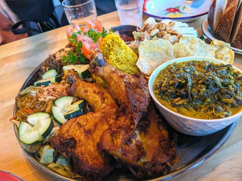 A plate filled with a variety of foods including grilled chicken, rice, vegetables, and a bowl of green leafy dish. A glass of water and another plate are in the background.
