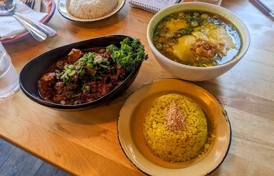 A meal with three dishes on a wooden table: a plate of seasoned yellow rice, a bowl of soup with green onions and meat, and a dish of stewed meat topped with herbs and green onions.