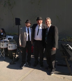 Three men in suits and hats stand together outdoors. Percussion instruments and a keyboard are visible in the background.