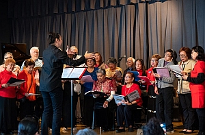A choir group, consisting mostly of older adults, sings on stage while being conducted by a leader dressed in black. Some members are standing, while others are seated. A blue curtain serves as the backdrop.