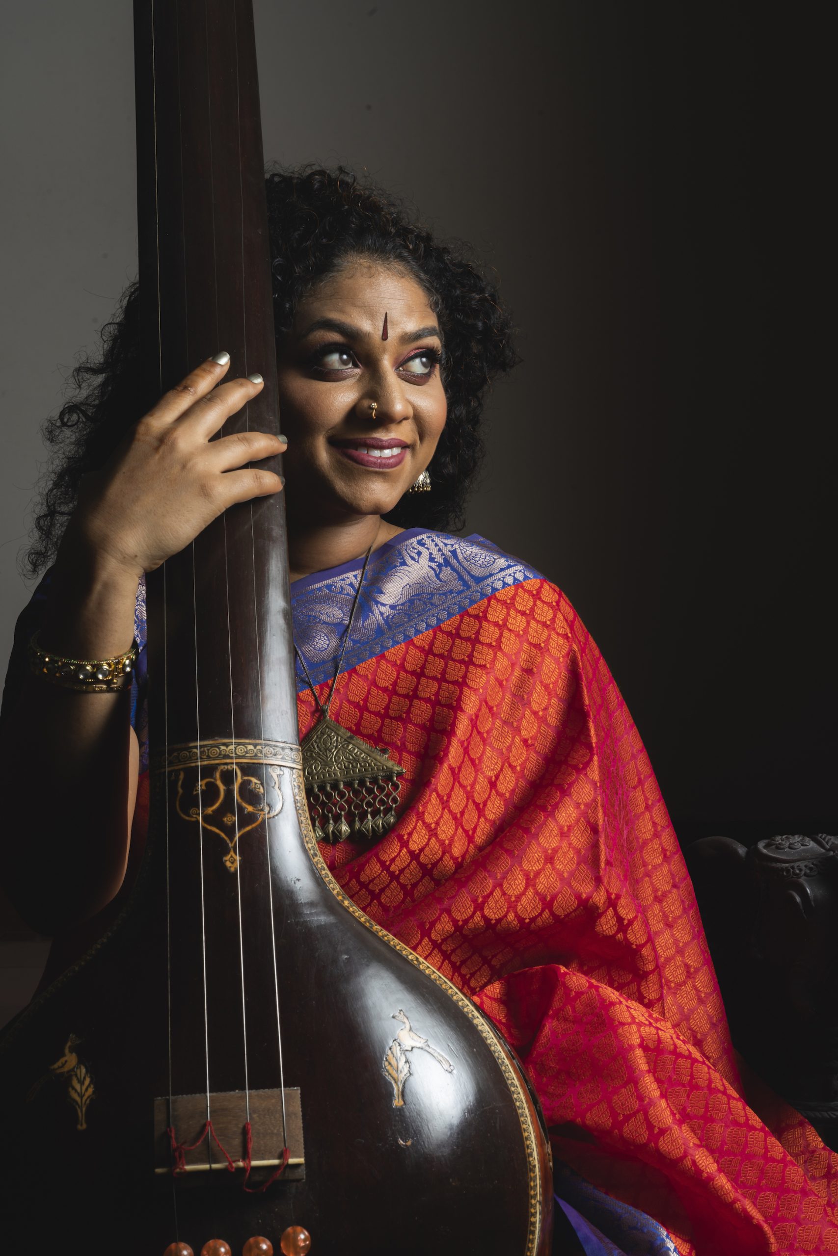 A person wearing a colorful saree holds a tanpura, an Indian classical string instrument. They are looking to the side, their expression reflective in the dimly lit setting, reminiscent of Sameer Gupta's artistic portraits.